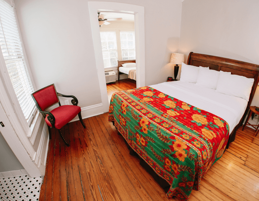 A cozy bedroom featuring a wooden bed with a colorful quilt, a vintage chair, and natural light from a window.