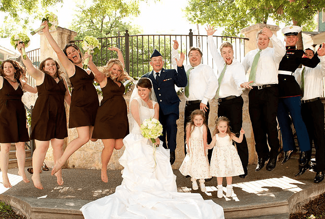 A joyful wedding party celebrates with arms raised, featuring the bride and groom in the center.