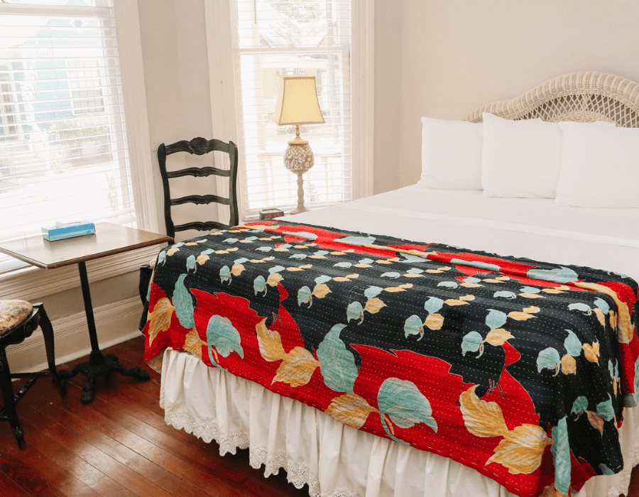 A cozy bedroom featuring a white bed with a colorful quilt, a small table, and a chair beside a window.