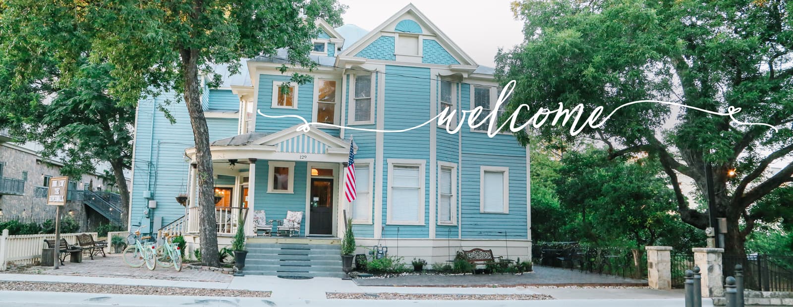 A blue Victorian-style house surrounded by greenery, featuring a welcoming sign.